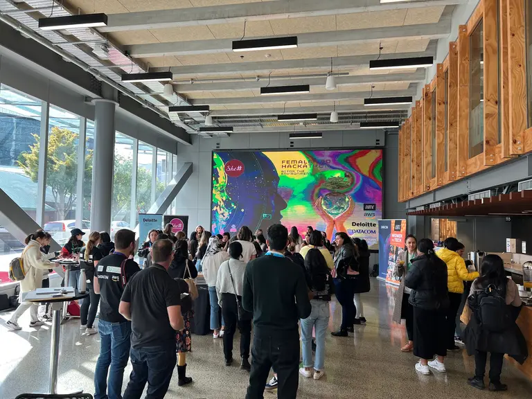 Attendees gathered in an event space beside a bright screen reading 'Female Led Hackathon' at the She Sharp hackathon.
