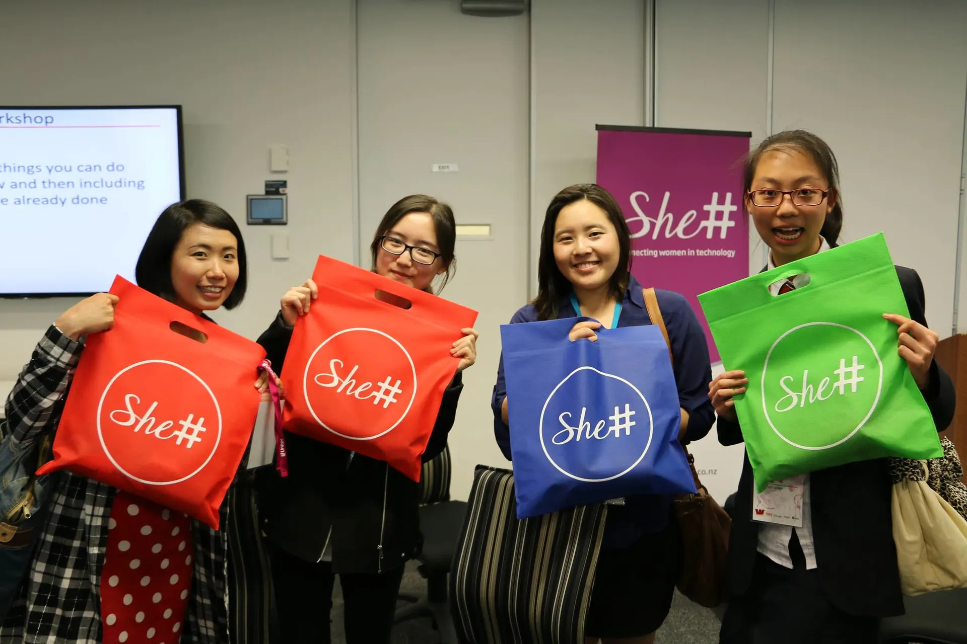 Attendees holding up colourful She Sharp tote bags at an event.
