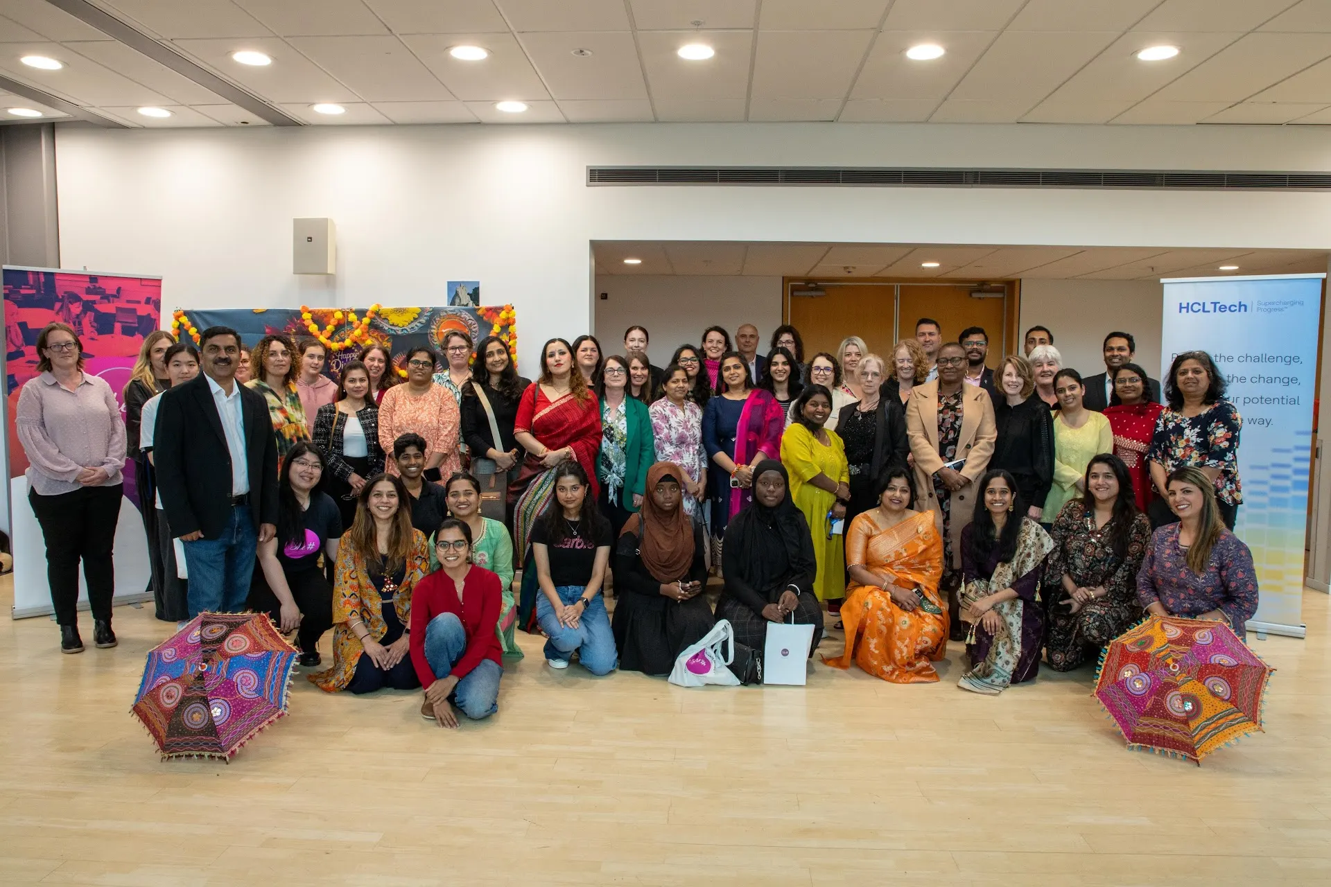 A large group in colourful traditional dress gathered for a group photo, with decorative parasols arranged in front, celebrating cultural diversity at a She Sharp event.