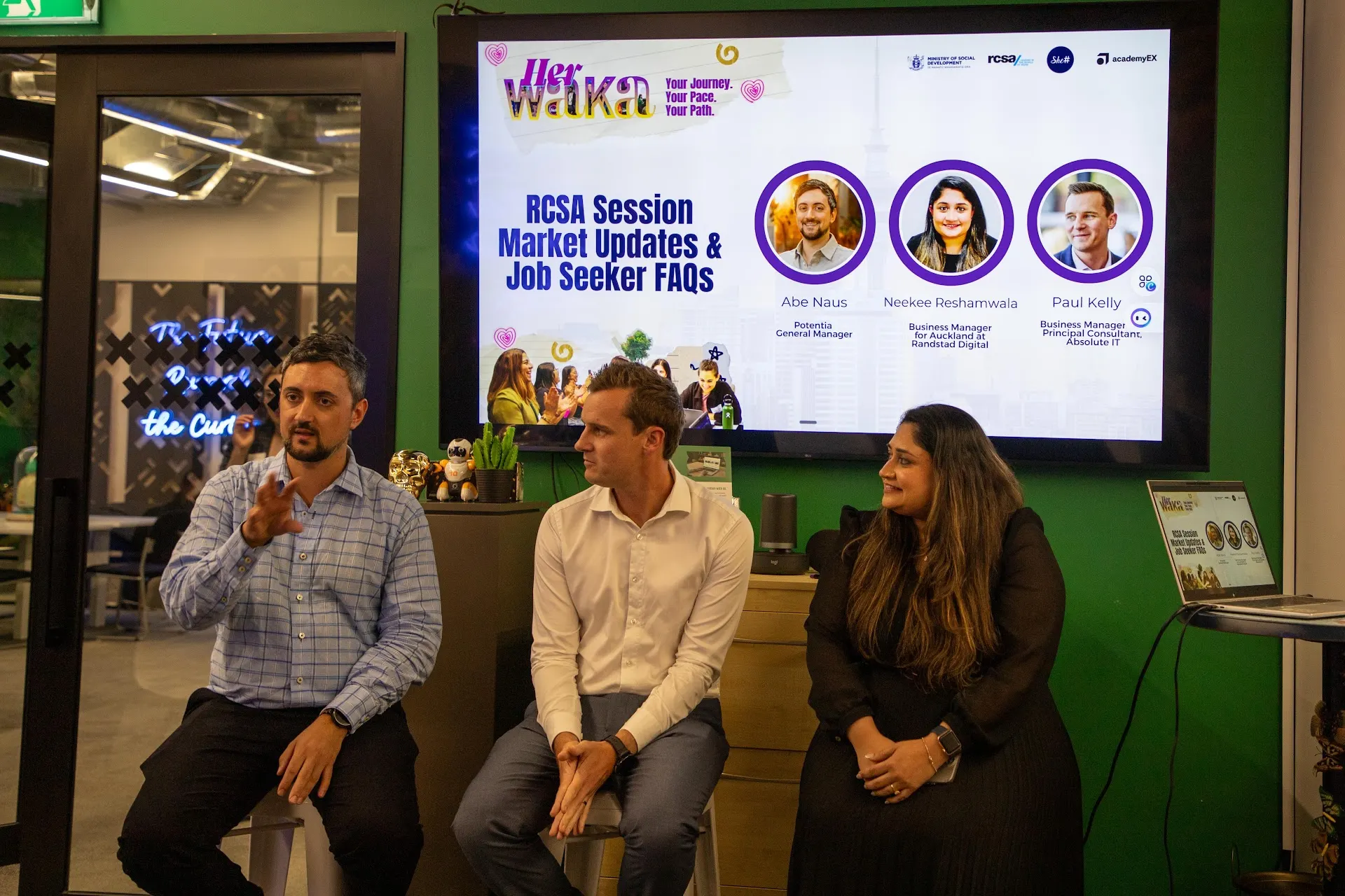 A panel of three speakers seated on stage during a She Sharp Her Waka session, with a presentation screen behind them.