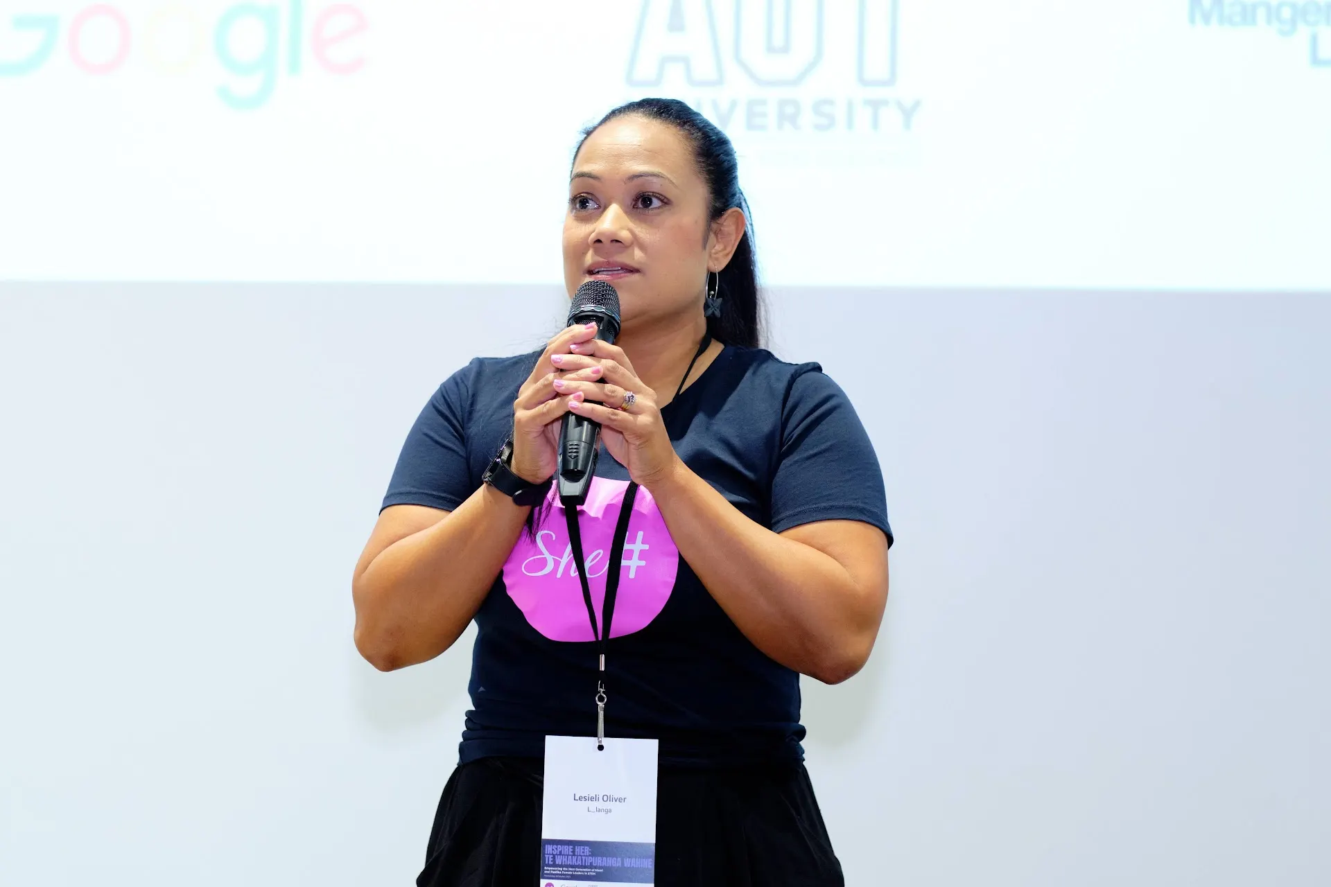 A speaker addressing the audience with a microphone at the She Sharp Inspire Her event, wearing a pink lanyard.